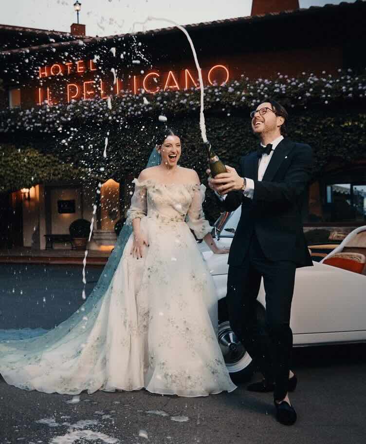 Newlywed couple popping a bottle of champagne in from of Hotel Il Pellicano.
